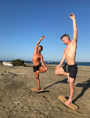 Zwei männliche Yogasportler balancieren auf einem Bein stehend auf einem Jungleboard am Strand.